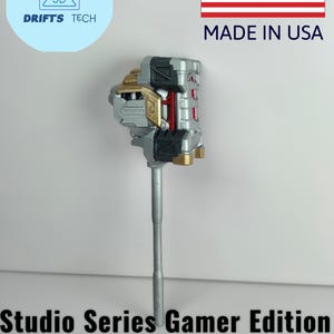 Puede incluir: Un martillo de juguete plateado, dorado y rojo, el Reactivate Gravity Maul de la serie Studio Gamer Edition. La imagen incluye el texto "Made in USA" y un logotipo que dice "Drifts Tech" y "3D".