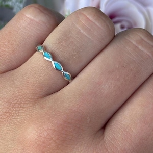 Op de afbeelding: Een zilveren ring met turquoise, marquise-vormige stenen. De ring zit om een hand, met een vage achtergrond van roze en witte bloemen. Het ontwerp van de ring heeft een herhalend patroon van de turquoise stenen.
