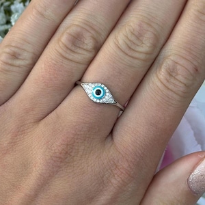 May include: A silver ring with an eye motif, a turquoise centre, and clear stone accents. The ring is displayed on a hand with manicured nails, set against a backdrop of white and pink flowers.