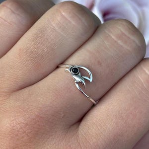 Puede incluir: Un anillo de plata con un diseño de media luna y una pequeña gema negra redonda en el centro. El anillo tiene una banda delgada y las puntas de la luna se curvan hacia arriba. El anillo se lleva en un dedo.