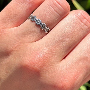 May include: A silver-colored ring featuring a delicate floral design. The ring is composed of small, detailed flowers arranged in a continuous pattern around the band. The ring is worn on a finger, with a blurred green background.