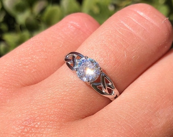 Könnte beinhalten: Ein silberner Ring mit einem keltischen Knotendesign und einem klaren, runden Edelstein in der Mitte.