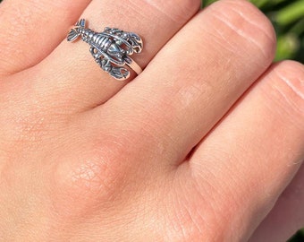 May include: A silver-colored ring featuring a detailed lobster design. The lobster is positioned horizontally across the finger, with its claws and body clearly visible. The ring is worn on a finger, showcasing the intricate craftsmanship of the jewelry.