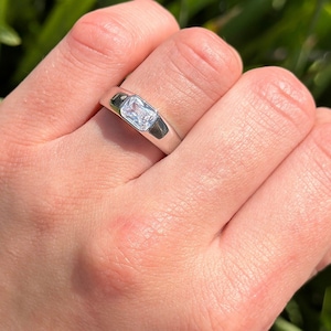 May include: A silver ring with a clear square-shaped gemstone. The ring is on a finger.