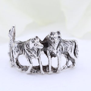 Puede incluir: Un anillo de plata con una escultura detallada de dos lobos de pie uno al lado del otro.
