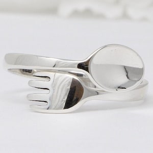May include: A silver ring shaped like a spoon and fork intertwined. The spoon is on top of the fork, and the spoon's bowl is facing the viewer.