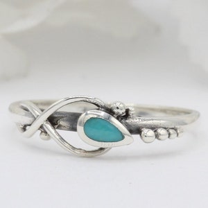 Op de afbeelding: Een zilveren ring met een turquoise traanvormige steen gezet in een gedraaide band. De band is versierd met kleine zilveren kralen.