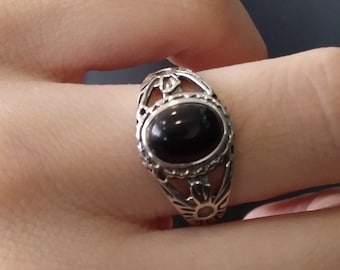 May include: A silver ring with an oval, black gemstone. The ring has an ornate, openwork design featuring sunburst and leaf-like details. The ring is shown on a finger.
