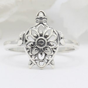 May include: A silver ring featuring a detailed, cutout design of a turtle with a flower-shaped shell.