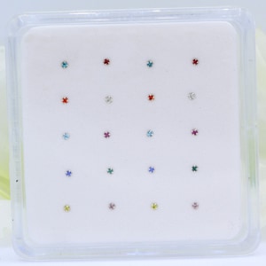 May include: A clear plastic box with 16 small silver nose rings with different colored gemstones. The gemstones are red, blue, green, yellow, and clear.