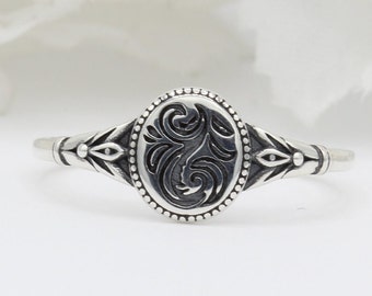 May include: A silver ring with an oval-shaped design featuring a detailed floral pattern. The ring has a beaded edge and a simple band.