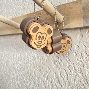May include: Three brown and tan plastic keychains featuring a raised, stylized cartoon mouse head design. Each keychain is attached to a silver-colored metal ball chain, allowing them to hang from a wooden peg.