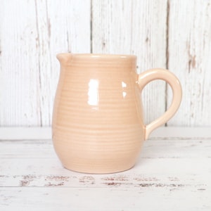 May include: A peach-colored ceramic pitcher with a curved handle and a spout. The pitcher has a smooth, glossy finish and a slightly textured surface. The background is a white wooden surface.
