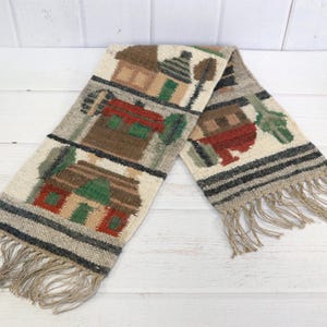 May include: Woven scarf featuring a repeating pattern of colorful houses and trees. The scarf has a cream-colored background with brown, red, green, and gray accents. The ends of the scarf have a fringe.