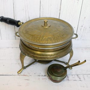 Puede incluir: Antiguo calienta platos de latón con tapa, asa y pequeño quemador. El calienta platos tiene un diseño detallado en la tapa y se apoya en tres patas ornamentadas. El asa es negra. Un pequeño quemador con tapa y pinzas está junto al plato.