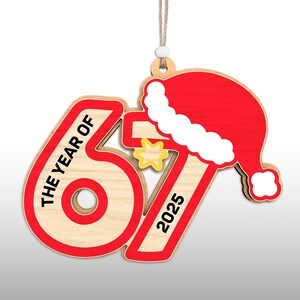 May include: Wooden Christmas ornament with the text "THE YEAR OF 67 2025" in red and black. The ornament features a red Santa hat and a yellow star. The ornament is hanging by a string.