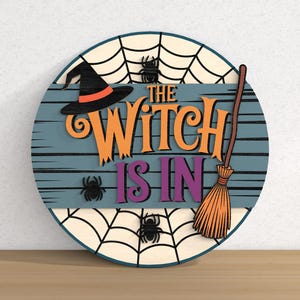 以下が含まれることがあります： 「THE WITCH IS IN」の文字が入った丸い木製のハロウィーンサイン。魔女の帽子、クモ、ほうき、クモの巣のデザインが特徴です。背景は青色で、水平線が入っています。