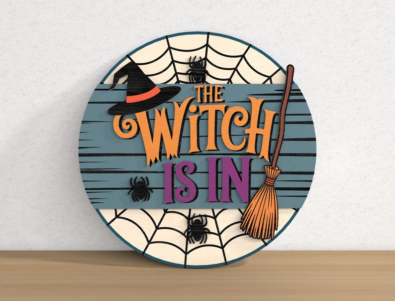 以下が含まれることがあります： 「THE WITCH IS IN」の文字が入った丸い木製のハロウィーンサイン。魔女の帽子、クモ、ほうき、クモの巣のデザインが特徴です。背景は青色で、水平線が入っています。