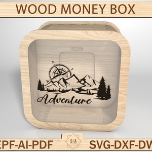 May include: A wooden money box with a clear front panel. The box has a compass, mountains, and trees engraved on the front. The text "Adventure" is also engraved on the front of the box.