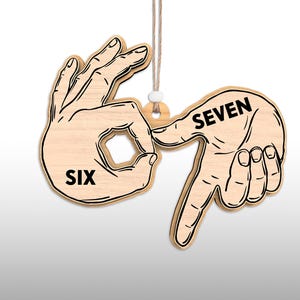 May include: Wooden ornament depicting the numbers six and seven in sign language. The word "SIX" is printed on one hand, and "SEVEN" on the other. The ornament is light brown with black outlines and hangs from a natural twine.