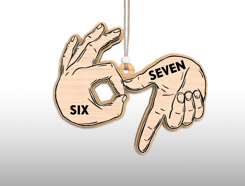 May include: Wooden ornament depicting the numbers six and seven in sign language. The word "SIX" is printed on one hand, and "SEVEN" on the other. The ornament is light brown with black outlines and hangs from a natural twine.