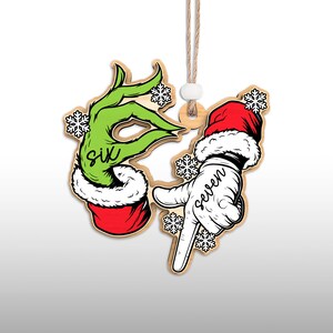 May include: Wooden Christmas ornament featuring two hands. One is green with the word "six" and the other is white with the word "seven". Both hands are wearing Santa hats and surrounded by snowflakes. The ornament hangs from a string.