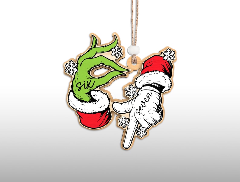 May include: Wooden Christmas ornament featuring two hands. One is green with the word "six" and the other is white with the word "seven". Both hands are wearing Santa hats and surrounded by snowflakes. The ornament hangs from a string.
