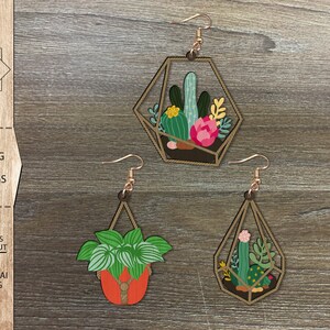 Op de afbeelding: Drie lasergesneden houten hangende planten oorbellen. De oorbellen hebben een geometrisch ontwerp met verschillende planten erin. De oorbellen zijn lichtbruin van kleur met een touwachtig ontwerp rond de randen.