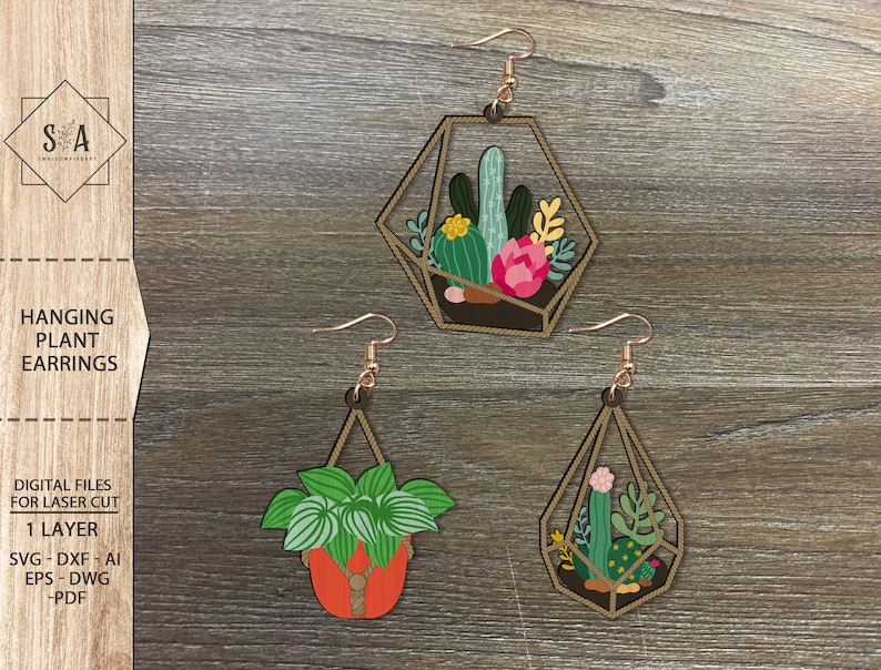 Op de afbeelding: Drie lasergesneden houten hangende planten oorbellen. De oorbellen hebben een geometrisch ontwerp met verschillende planten erin. De oorbellen zijn lichtbruin van kleur met een touwachtig ontwerp rond de randen.