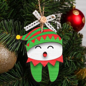 May include: A green and red wooden Christmas ornament shaped like a tooth with a smiling face and a green elf hat. The ornament has a white background and a red mouth.