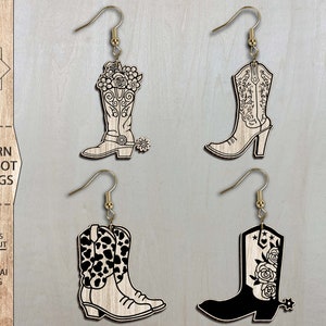 May include: Four pairs of laser cut wooden earrings shaped like cowboy boots. The boots have different designs, including floral, cow print, and a solid black boot with floral accents.