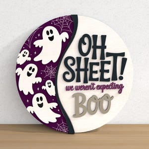 以下が含まれることがあります： ハロウィーンをテーマにした丸い木製の看板。看板は、黒で「OH SHEET!」、紫で「we weren't expecting」、グレーで「BOO」というテキストが書かれた白い背景が特徴です。白い幽霊とクモの巣が付いた紫色の三日月が左側にあります。
