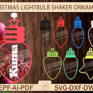 Può includere: Un ornamento natalizio a forma di lampadina rossa con un elemento shaker. L'ornamento è decorato con glitter bianco e cuori rossi. L'ornamento è etichettato "Kuma". L'immagine mostra anche diversi altri ornamenti a forma di lampadina in diversi colori, tra cui rosso, blu, verde e giallo.