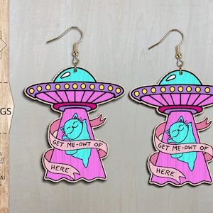 Può includere: Un paio di orecchini a forma di UFO rosa e blu con un gatto all'interno. Gli UFO hanno cerchi gialli intorno ai bordi. Il gatto è blu e ha il testo "Get me-owt of here" su uno stendardo rosa.