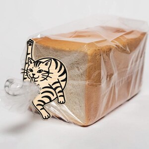 May include: A loaf of sliced bread in a clear plastic bag, secured with a silver clip and a decorative wooden cat-shaped tag. The cat has black stripes and is reaching up with one paw.