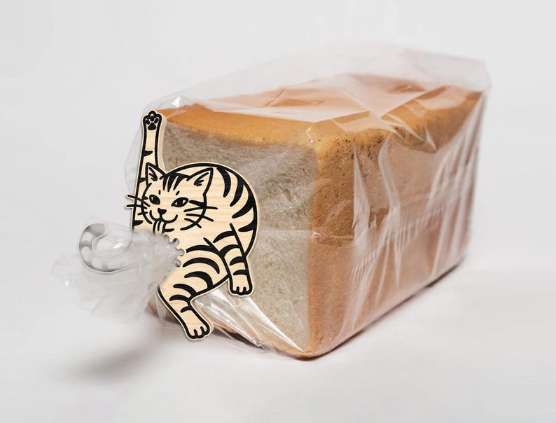 May include: A loaf of sliced bread in a clear plastic bag, secured with a silver clip and a decorative wooden cat-shaped tag. The cat has black stripes and is reaching up with one paw.
