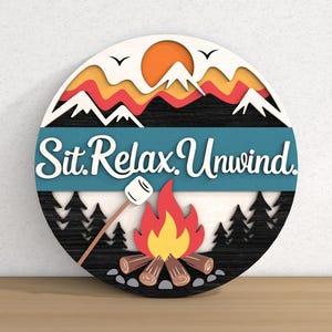 May include: A round wooden sign with a camping theme. The sign features a campfire, mountains, and the words "Sit. Relax. Unwind." in white script against a teal banner. The design includes a marshmallow on a stick and a rising sun.
