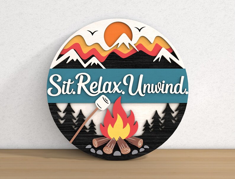 May include: A round wooden sign with a camping theme. The sign features a campfire, mountains, and the words "Sit. Relax. Unwind." in white script against a teal banner. The design includes a marshmallow on a stick and a rising sun.