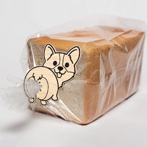 May include: A loaf of sliced bread in a clear plastic bag, secured with a wooden corgi-shaped bread clip. The clip is light brown with black details, depicting the dog's face and rear.