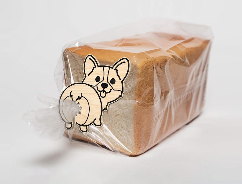 May include: A loaf of sliced bread in a clear plastic bag, secured with a wooden corgi-shaped bread clip. The clip is light brown with black details, depicting the dog's face and rear.