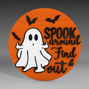 Op de afbeelding: Een rond, oranje houten bord met een witte geest met zwarte ogen en een zwarte mond. De geest steekt zijn middelvinger op. Het bord heeft ook zwarte vleermuizen en de tekst "SPOOK around Find out &" in zwart.