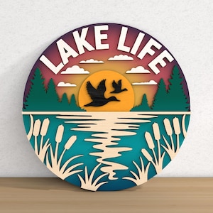 May include: A round, layered wooden sign with a lake scene. The sign features the words "LAKE LIFE" in white, a sunset, flying birds, trees, and water. The color palette includes shades of blue, green, orange, and purple.