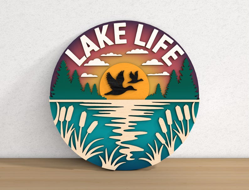 May include: A round, layered wooden sign with a lake scene. The sign features the words "LAKE LIFE" in white, a sunset, flying birds, trees, and water. The color palette includes shades of blue, green, orange, and purple.