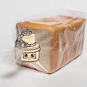 May include: A loaf of sliced bread in a clear plastic bag. A wooden cake-shaped tag with a floral design and a cartoon face is attached to the bag. The bread has a golden-brown crust and a soft, white interior.