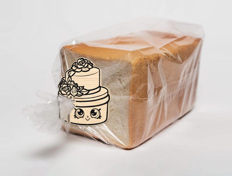 May include: A loaf of sliced bread in a clear plastic bag. A wooden cake-shaped tag with a floral design and a cartoon face is attached to the bag. The bread has a golden-brown crust and a soft, white interior.