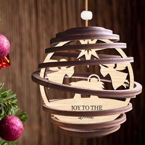 Puede incluir: Un adorno navide&ntilde;o de madera con un dise&ntilde;o en capas que representa una escena de la natividad. El adorno es marr&oacute;n y tiene la inscripci&oacute;n "Joy to the World".