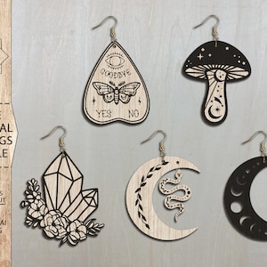May include: A set of four laser cut wooden earrings featuring mystical designs. The earrings include a crescent moon with a snake, a crystal, a mushroom with stars, and a Ouija board with a butterfly.