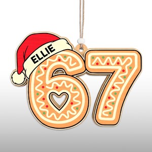 May include: A wooden Christmas ornament shaped like the numbers 6 and 7, decorated to resemble gingerbread cookies. The number 6 has a heart-shaped cutout and is topped with a red Santa hat with the name "ELLIE". The ornament hangs from a string.