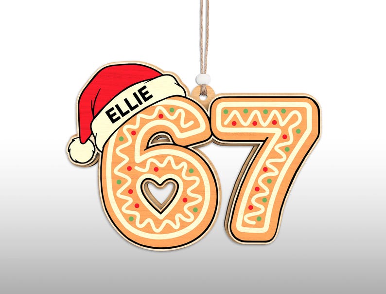 May include: A wooden Christmas ornament shaped like the numbers 6 and 7, decorated to resemble gingerbread cookies. The number 6 has a heart-shaped cutout and is topped with a red Santa hat with the name "ELLIE". The ornament hangs from a string.
