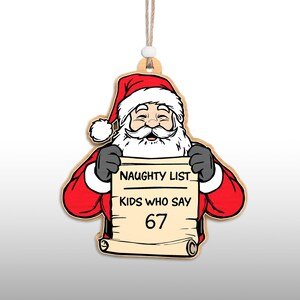 May include: Wooden Christmas ornament featuring Santa Claus holding a scroll. Santa is dressed in a red suit and hat with a white beard. The scroll reads "NAUGHTY LIST KIDS WHO SAY 67". The ornament has a string for hanging.